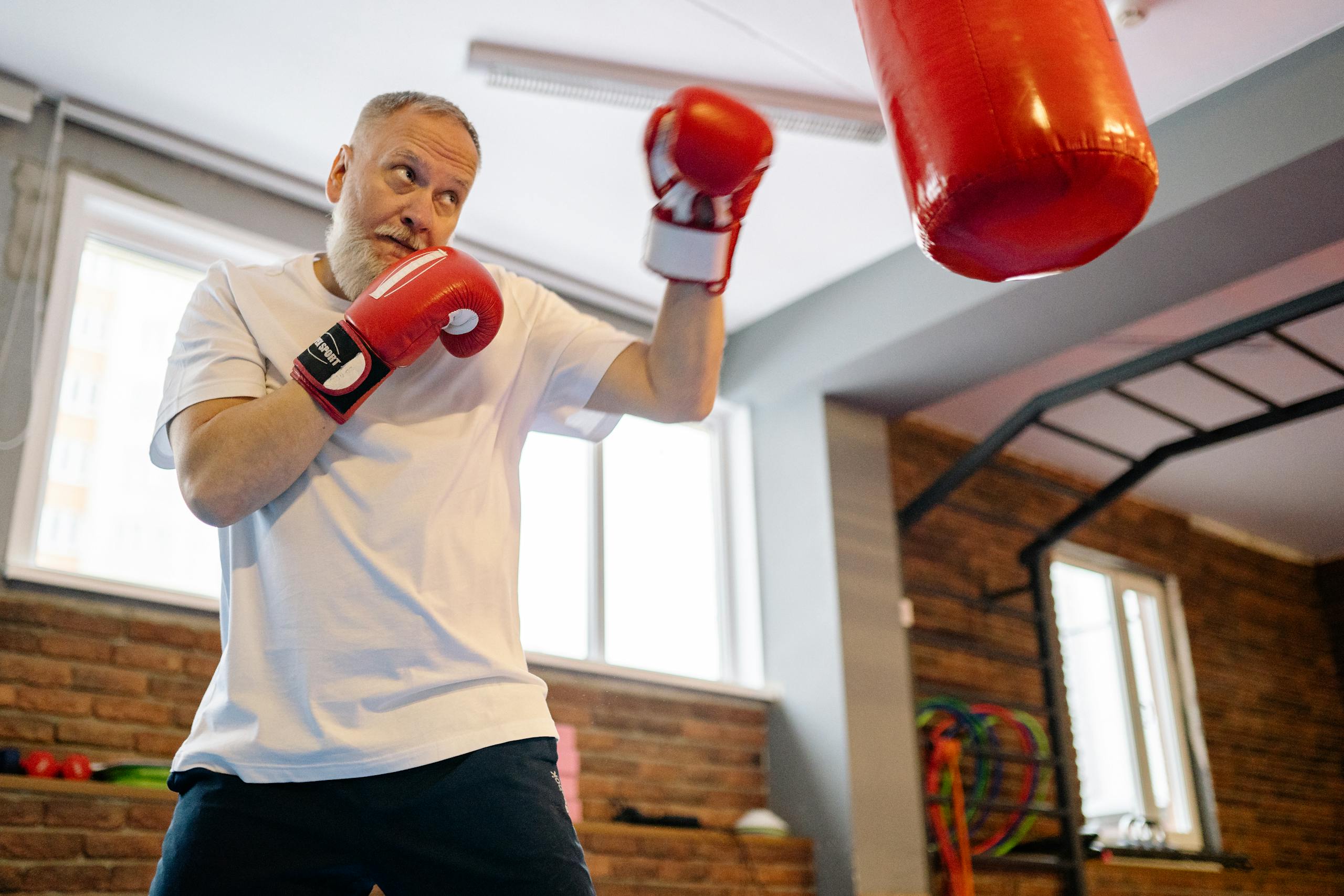 Elderly man practicing boxing indoors with gloves and punching bag.