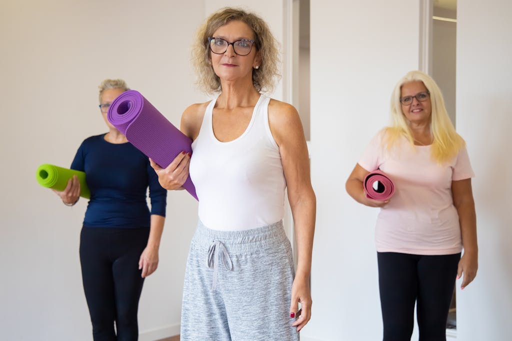 Three senior women holding yoga mats, preparing for a yoga class indoors.