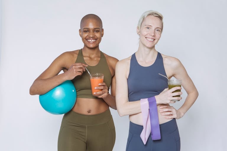 Two diverse women smiling, holding smoothies and fitness gear, promoting health and wellness.